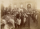 Photograph of crowd in front of Harrison's North Delaware Street home in 1888. Benjamin Harrison is standing near the front door slighlty to the left. Image shows the front of the house as it looked in 1888, picket fence at front sidewalk, and crowd of mostly men and young boys. A few women stand behind Harrison at the front door. The crowd is lined up on the rightside of the walk leading to the front door, in the yard to the left, and on the front sidewalk to the left.