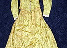 Gold flowered silk one piece dress marked as being Caroline Harrison's. Fabric is a gold color and different shades outlined in black of a design of long stemmed flowers. Like a poppy or daisy. Lace trimmed collar and cuffs. Lined, bottom edge of skirt inside small ruffle of a stiff netting. 13 buttons on front of bodice. Bodice attached at back with skirt. Metal stays some coming through fabric.