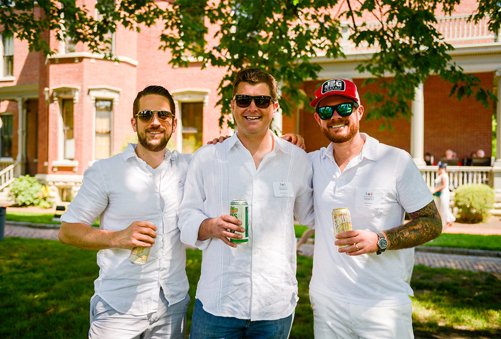 Photograph of a trio of males having a good time at the Presidential Site, all dressed in white, sporting sunglasses, holding drink cans, and relaxing in the shade of an oak tree in the side yard.