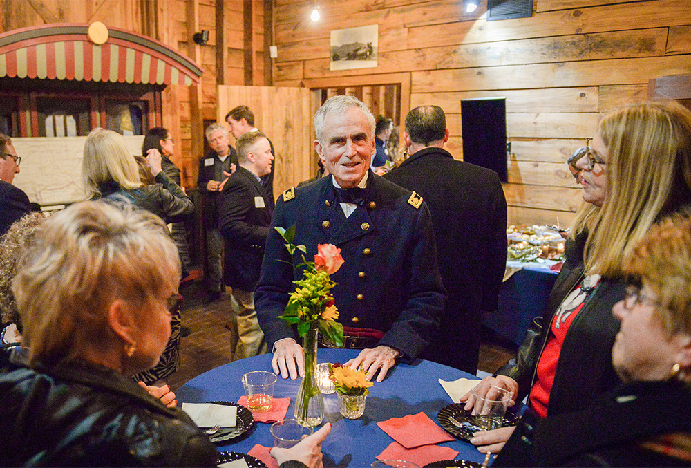 Photograph of a union soldier standing in the presidential site welcome center, speaking to a small crowd of people at a dinner table.