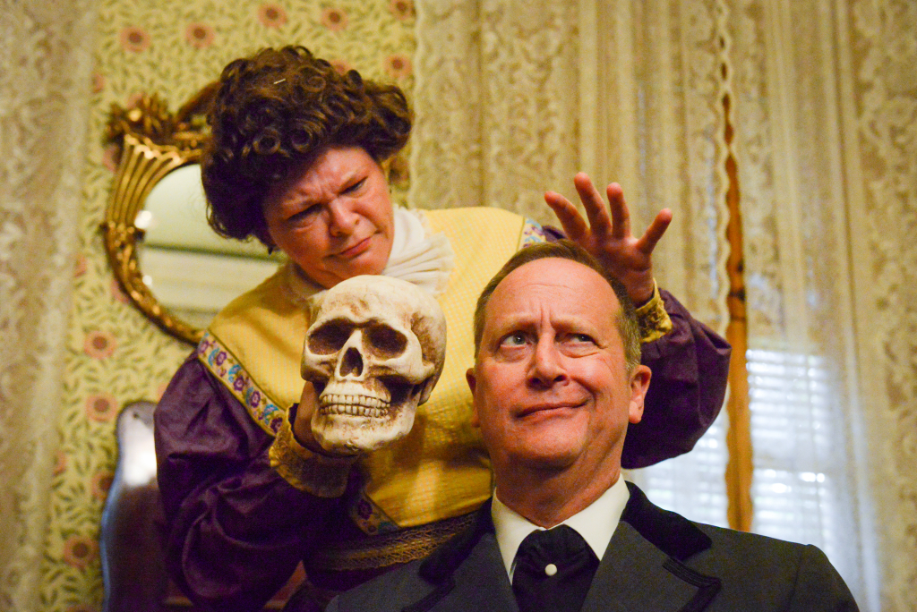 photo of two comedies of error actors. a woman is standing on the left, holding a human skull and holding a hand above a seated man's head while he has a curious expression on his face.