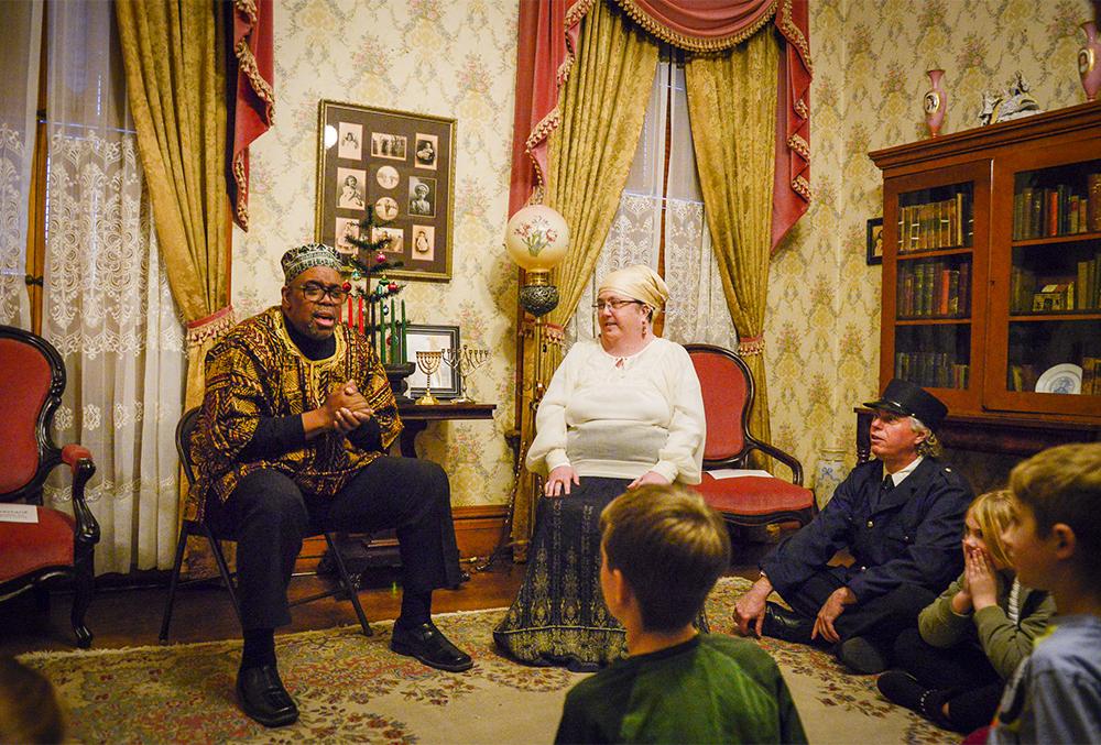 Photograph of two candlelight theater actors, one playing befana and the other maggid. they are seated in a room of the presidential home as a crowd of onlookers views their performance.