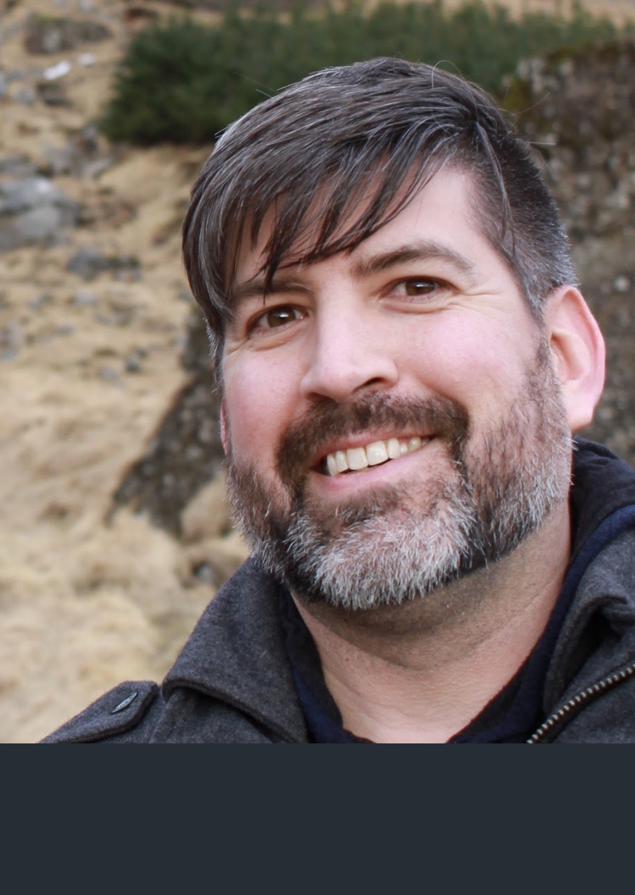Photograph of Brendan O Sullivan Hale, a man in his middle ages with a dark jacket, dark greying beared, and dark medium length hair. He has brown eyes and smiles at the camera.