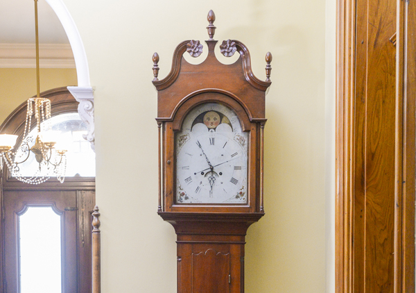 Tall case or grandfather clock made by George Woltz in Hagerstown, Maryland between 1800 and 1812. Clock has moon dial at top and a date dial below the center of the hands, and strikes on the hour. Cherry cabinet with front legs that curve out . There is a spindle on either side of the face. Rounded center at the top with rosette design. Top corners come up to pointed spindles. Clock face is a half circle at top with the dates above the moon dial, Roman numerals and painted floral designs in four corners, and glass door.