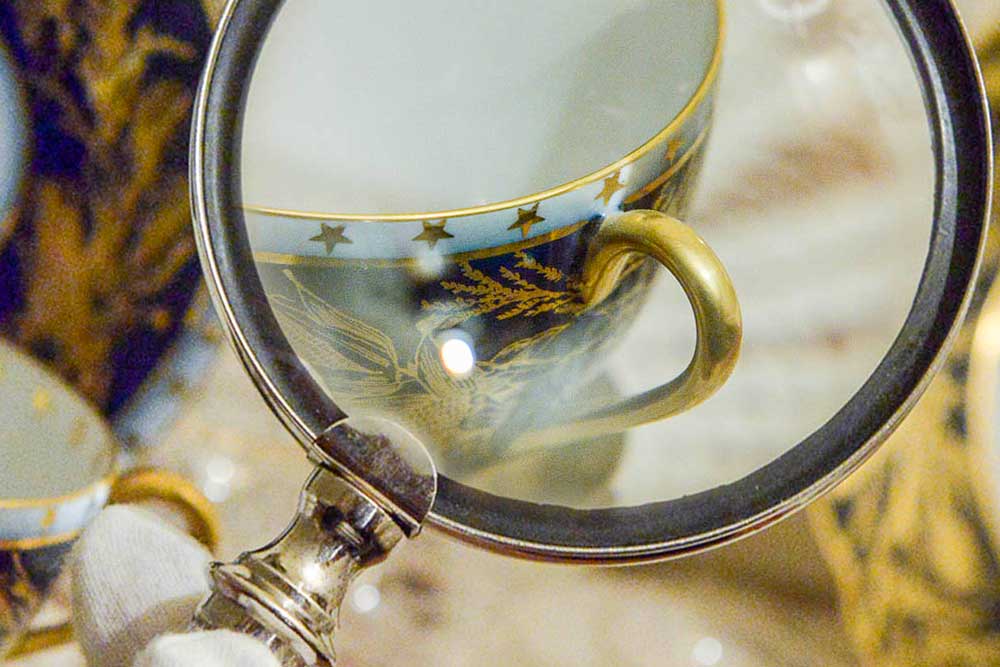 Photograph of a magnifying glass enhancing the detail on a coffee cup. The cup is inlaid with gold details depicting tree branches and stars, as well as a gold trim around the lip of the cup and handle.