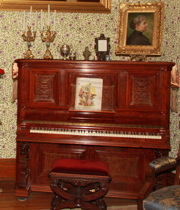 Wooden upright piano. There is a silver plaque mounted above the keyboard with manufacturer's name and information. The wood panels on each side of front have an incised design. Center panel has incised design of violin and mandolin crossed surrounded by flowers and leaves.