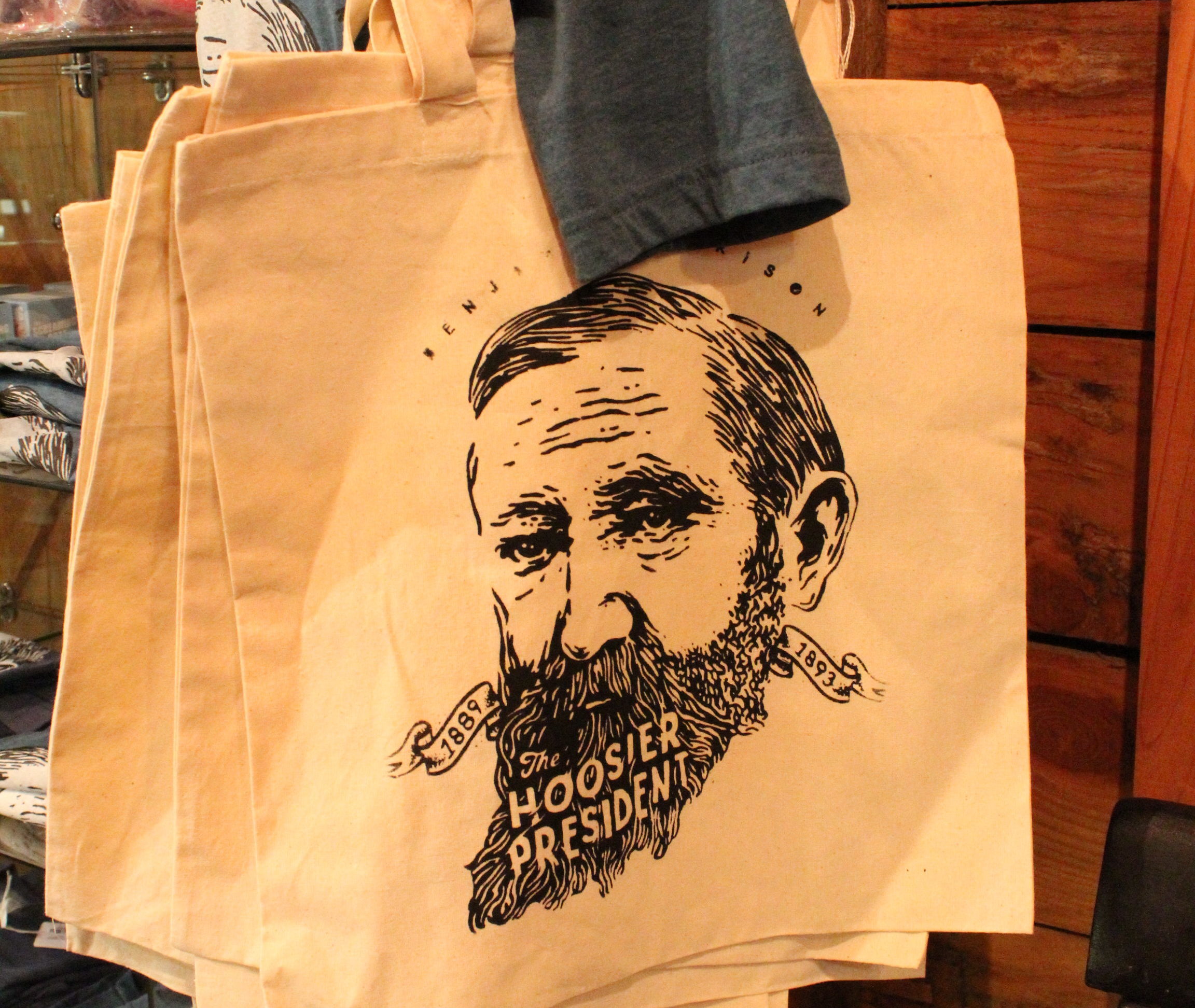 A photograph featuring an tote bag sold at the presidential site gift shop, emblazoned with an artful illustration of Benjamin Harrison's face. There is text in the negative space in his beard, reading "The hoosier president".