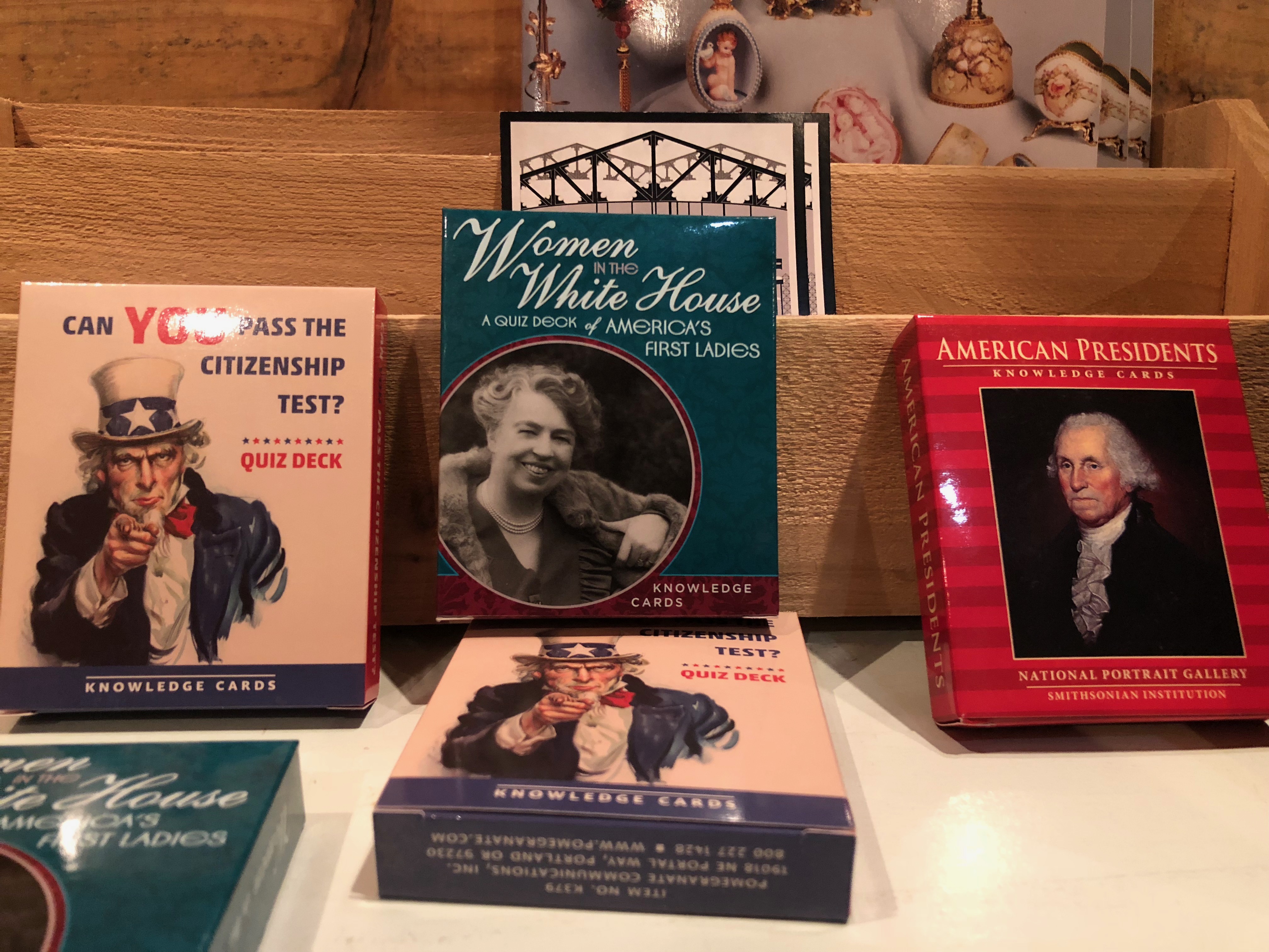 Photograph of several packs of knowledge cards sold at the Benjamin Harrison gift shop. Pictured from left to right: "Can you pass the citizen ship test? Quiz deck. The cover art features the famous image of uncle sam pointing at the viewer." "Women in the White House: A quiz deck of America's first ladies", featuring a photo of Eleanor Roosevelt. Lastly pictured is a red stripped book featuring a portrait of George Washington. The title reads "American Presidents, knowledge cards. National portrait gallery, smithsonian institution.