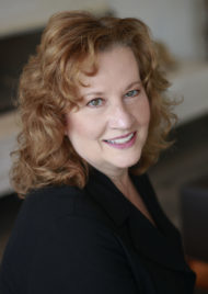 Portrait of Anne Surina. She is middle aged with reddish brown hair and green eyes. She smiles at the camera and wears a black dress jacket.