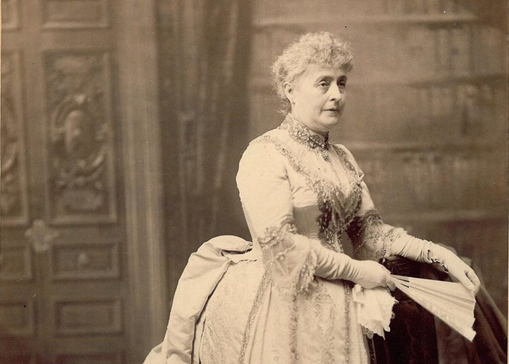 Sepia-toned photographic image of Caroline Harrison in her inaugural dress which has a large bustle and beading on the bodice and around the neck. Caroline is wearing gloves and holding a fan; her hair is pulled back from her face. She is standing in front of a large bookcase and is resting her hands in the back of a chair, while holding a fan and kerchief.