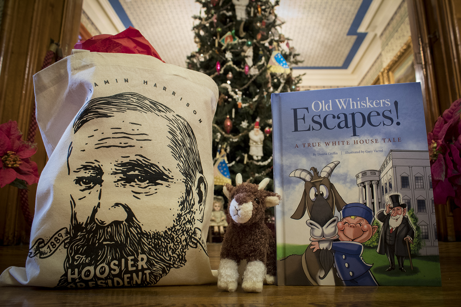This photograph features Benjamin Harrison merchandise, including a tote bag sporting an illustration of Benjamin Harrison's face, a plush of Benjamin Harrison's pet goat, Old Whiskers, and a children's book about Old Whisker's escape. Pictured in front of a holiday tree, fully decorated with ornaments and tinsel.