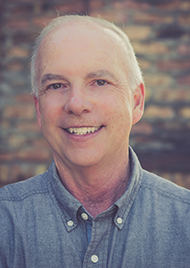 Portrait of James Hayes, an elderly man with short white hair and dark eyes. He wears a light blue collared shirt and grins at the camera.