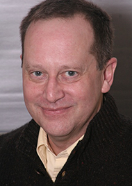 Portrait of Ken Eder, a middle aged man with dark brown hair and dark eyes. He wears a black jacket with lightly colored undershirt, and a smile.