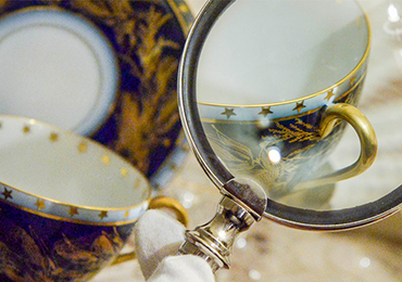 A photograph of an intricate coffee cup, complete with gold lining, gold handle, and stars along the mouth of the cup. There is a magnifying glass being used to enhance the detail.