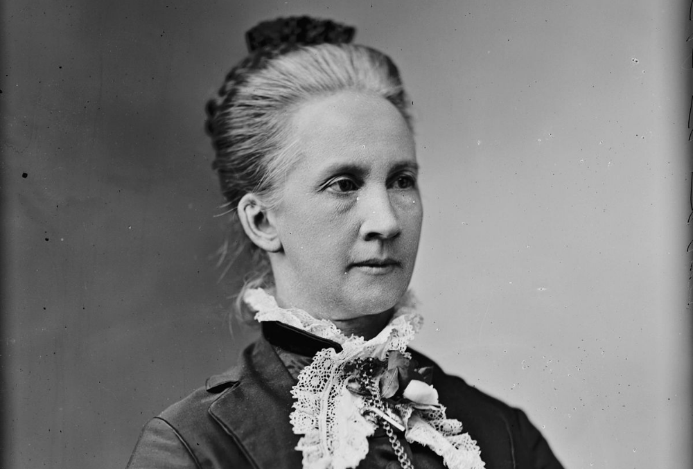 Photo of Belva Lockwood, dressed darkly with small white frills at the neck. Her hair is loosely bunned and braided with a black hairpin. She intensely looks out of frame, to the right of the photographer.